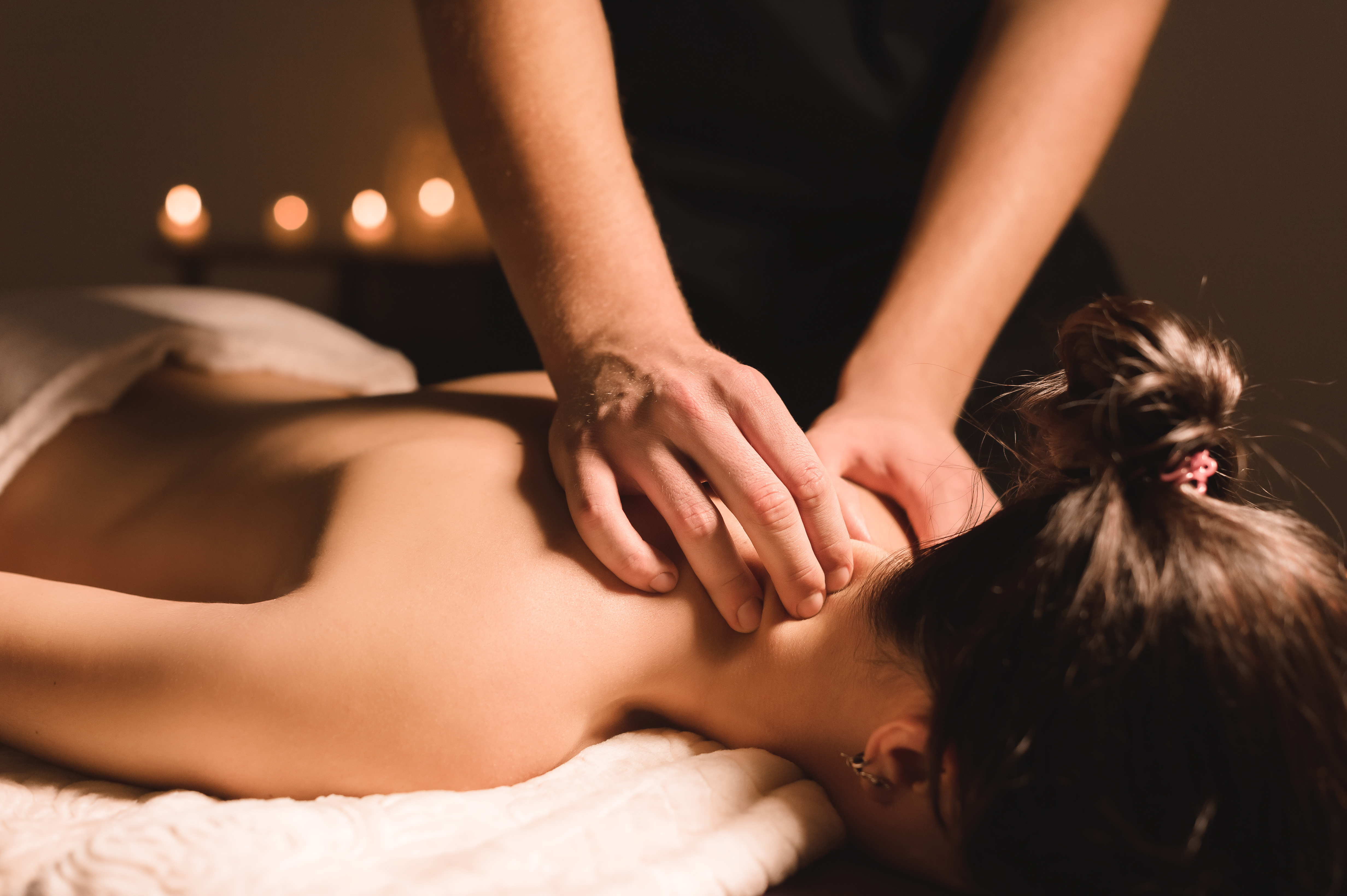 Men's hands make a therapeutic neck massage for a girl lying on a massage couch in a massage spa with dark lighting. Close-up. Dark Key